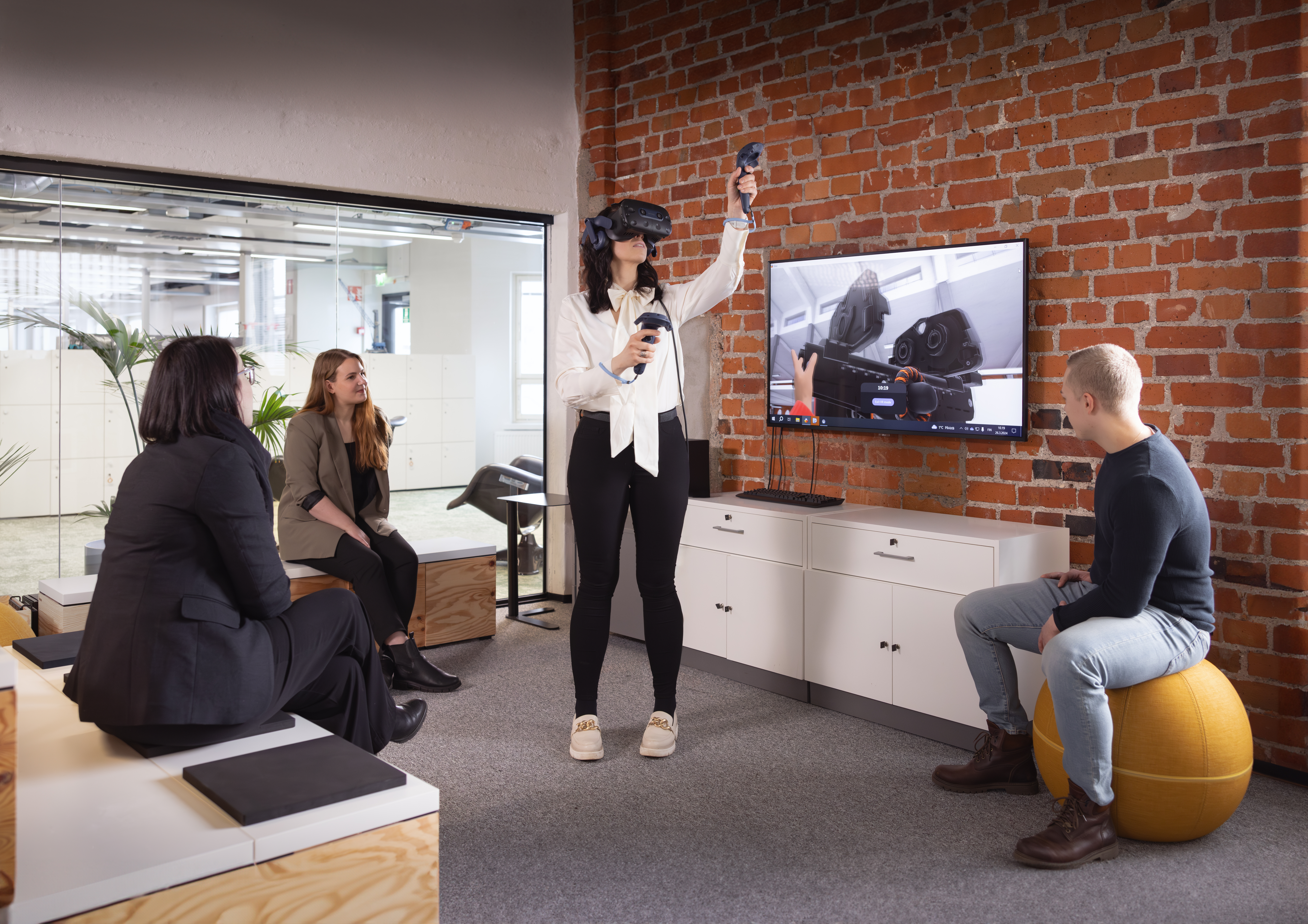 Drei Personen sitzen in einem Büro um einen Fernseher herum, während eine Frau mit VR-Brille und Controllern in der Mitte vor dem Fernseher steht.